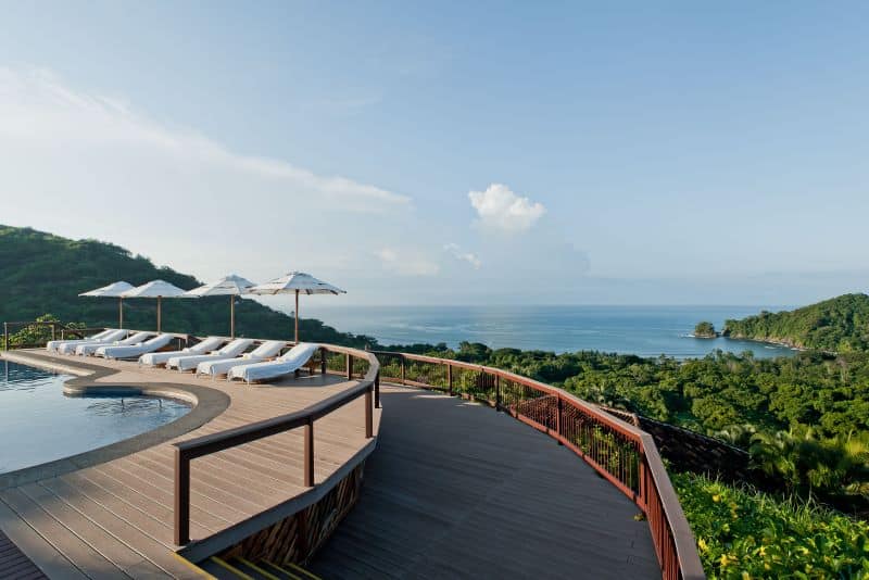 Pool deck with white lounge chairs and sun umbrellas overlooking ocean and green hills