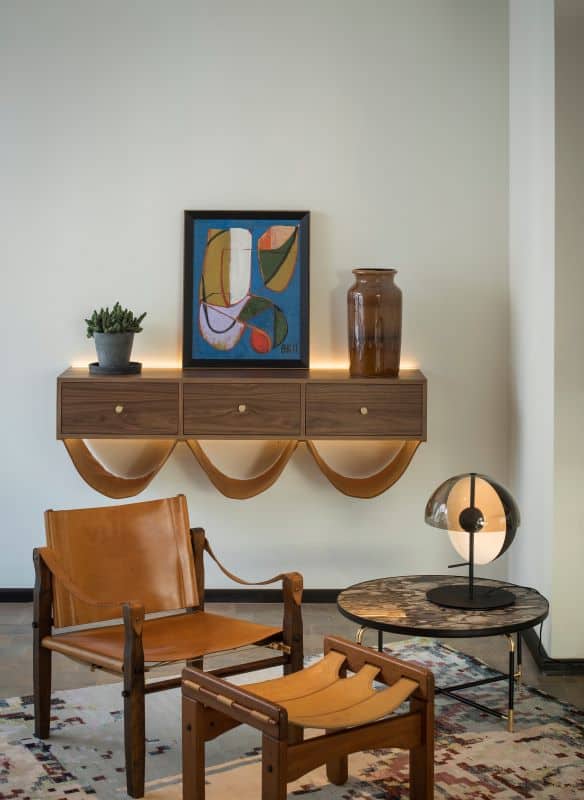 Wooden framed leather chair and footrest in front of shelf of drawers with artwork and vase on top